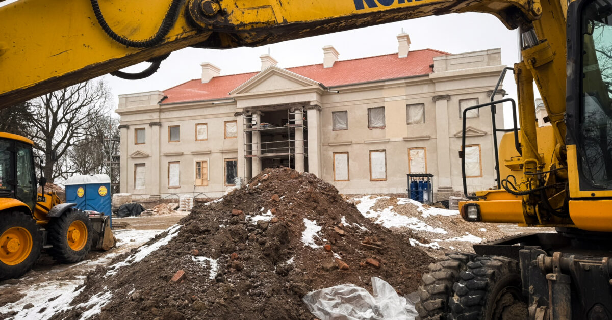 Zdjęcie przedstawiające plac budowy w Nawrze; na pierwszym planie znajduje się wysięgnik koparki i usypana ziemia, w głębi widać remontowany klasycystyczny pałac w Nawrze