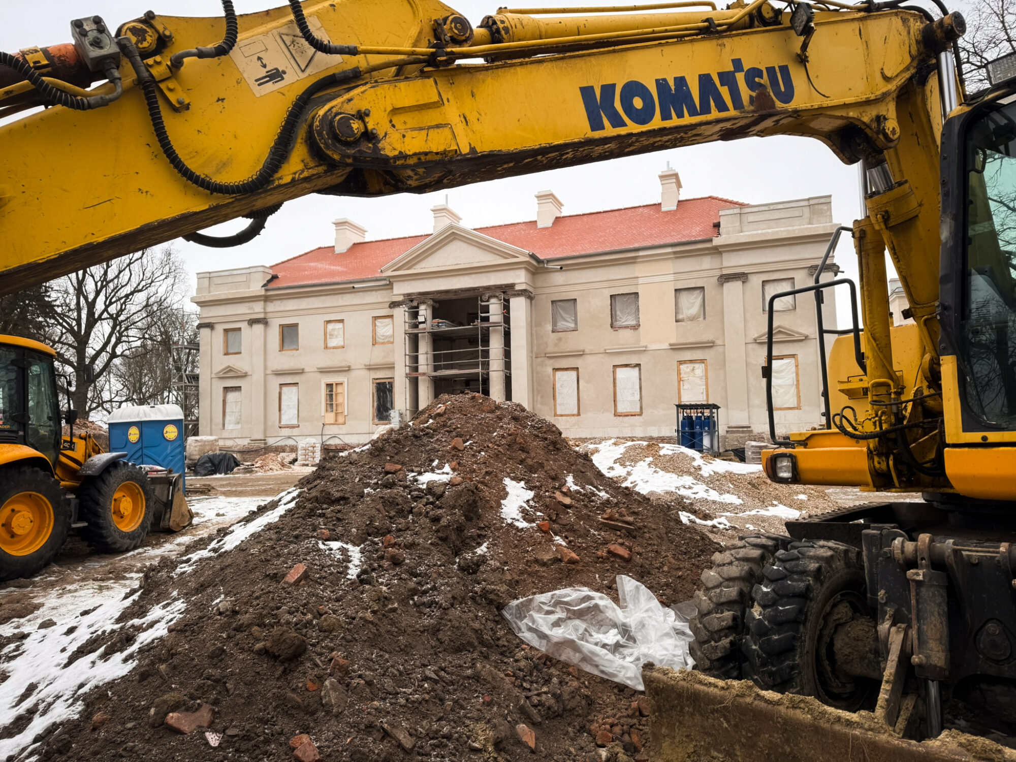 Zdjęcie przedstawiające plac budowy w Nawrze; na pierwszym planie znajduje się wysięgnik koparki i usypana ziemia, w głębi widać remontowany klasycystyczny pałac w Nawrze