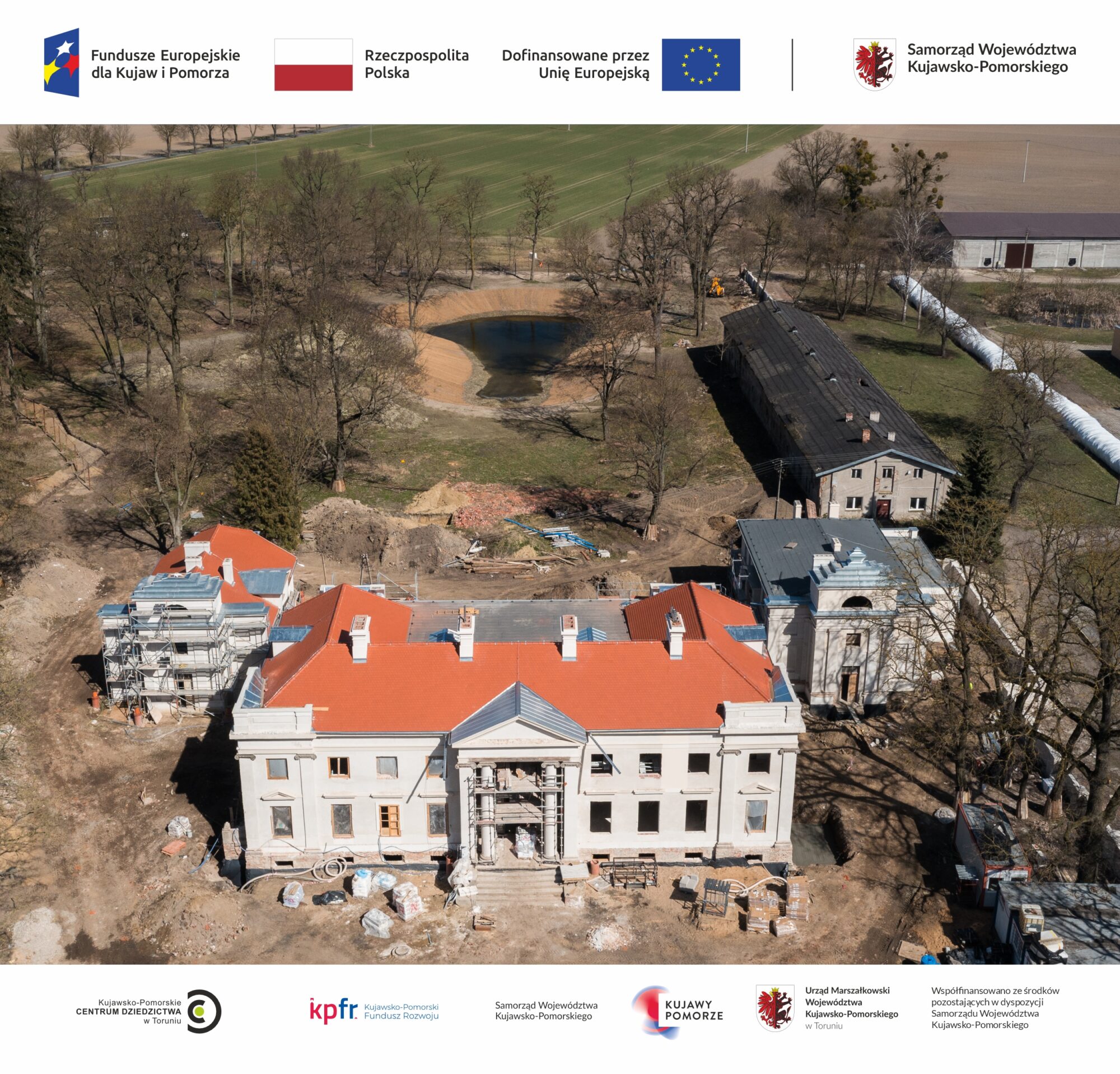 Zdjęcie przedstawiające plac budowy w Nawrze; na pierwszym planie znajduje się klasycystyczny pałac w Nawrze z jasną elewacją i dachem pokrytym czerwoną dachówką; po jego bokach dwie oficyny; w głębi staw otoczony drzewami i stara chlewnia